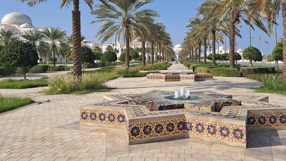 A mosaic-tiled fountain on a palm-lined path leading to the white domes of Qasr Al Watan