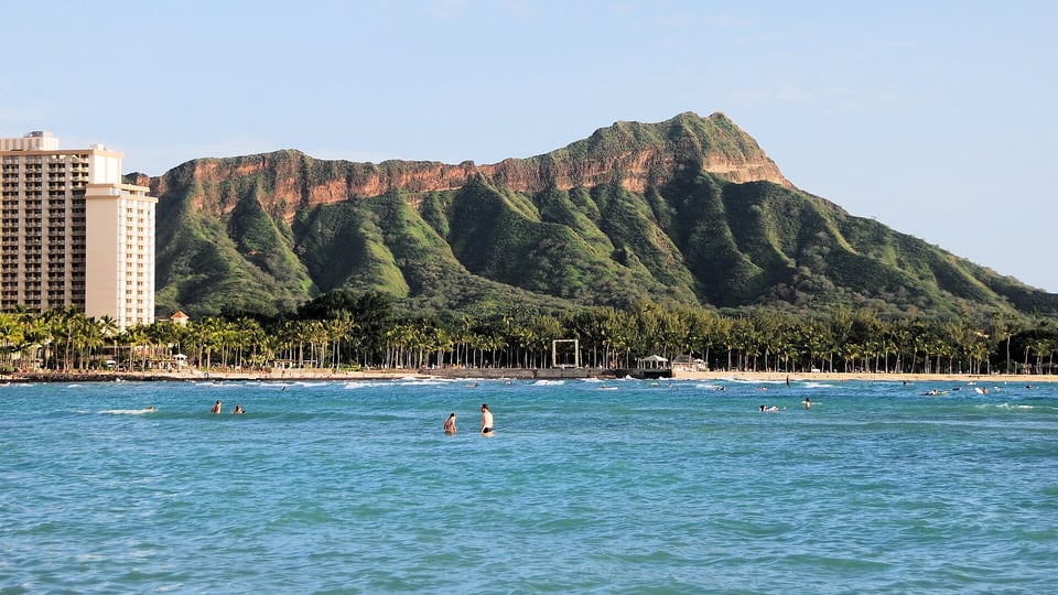 Diamond Head rising across the water off Waikiki Beach, with swimmers offshore and a hotel tower at the left