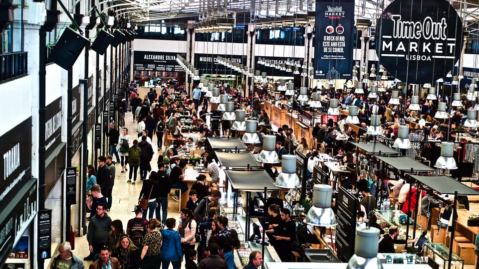 Diners at shared tables beneath the Time Out Market sign inside Mercado da Ribeira, Lisbon