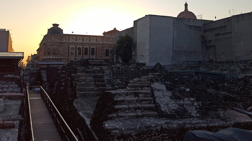 Stepped Templo Mayor foundations at dusk with colonial buildings behind them in the Centro Histórico