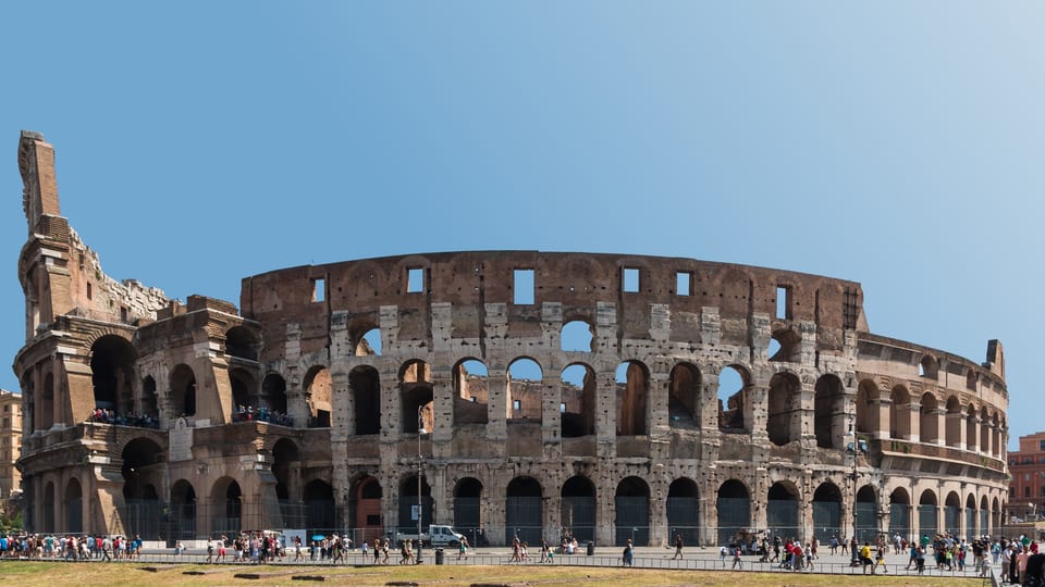 The outer walls and arches of the Colosseum in Rome under a clear sky, with visitors along its base
