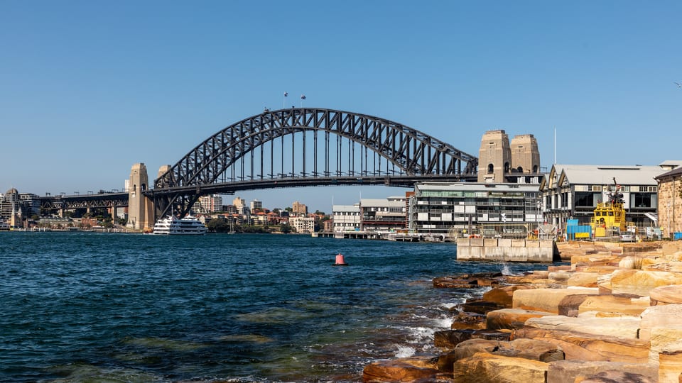 The steel arch of the Sydney Harbour Bridge seen across the water from the sandstone foreshore